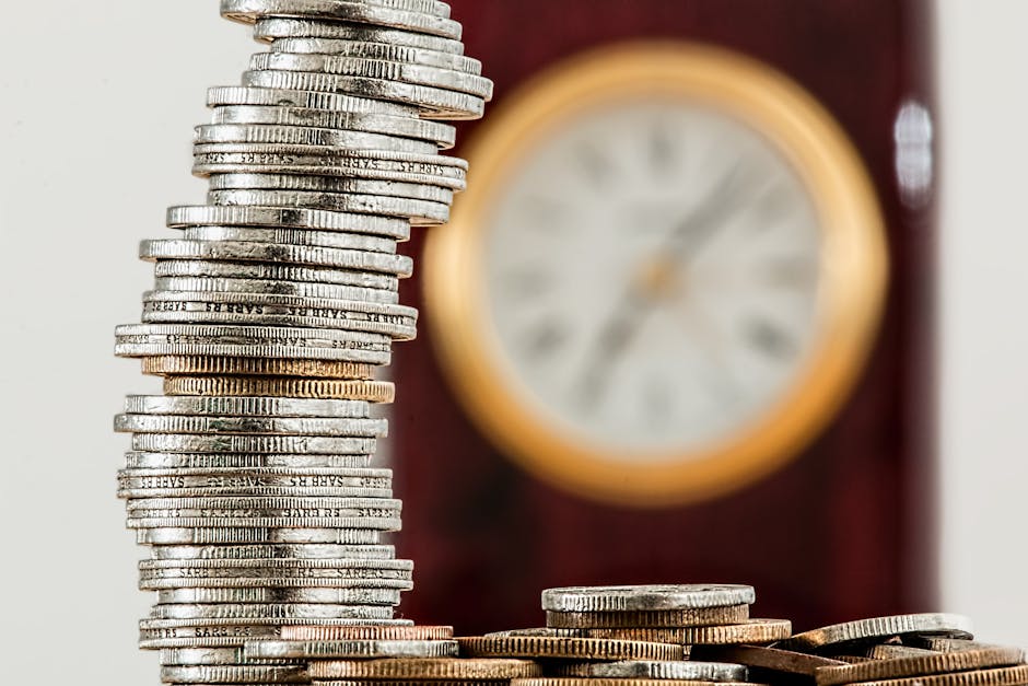 Close-up view of a tall, uneven stack of assorted UK coins, including silver and gold-colored coins, placed on a flat surface inside a home. In the background, a wooden antique clock with an off-white