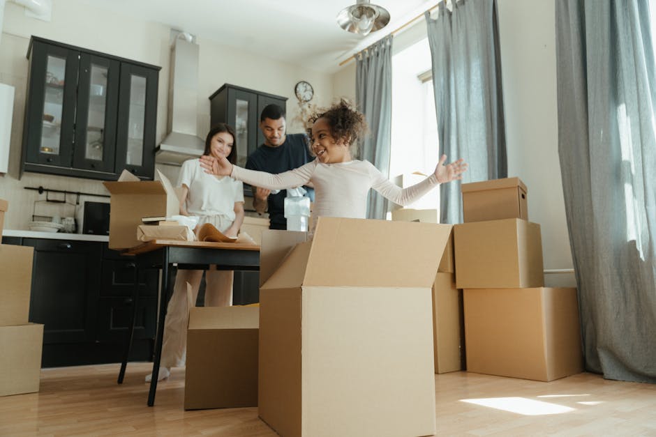 A family of three preparing for a home relocation inside a well-lit living room, with large windows and blue curtains. The mother and father stand behind a black table that has several open cardboard 