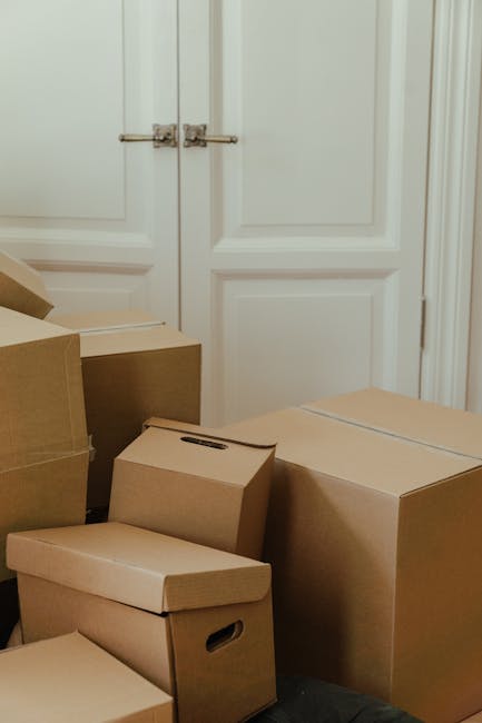 Close-up of a person wearing a white shirt, holding a wrapped glass or fragile item covered in bubble wrap, while preparing to place it into a cardboard box labeled 'KITCHEN' in a domestic kitchen set
